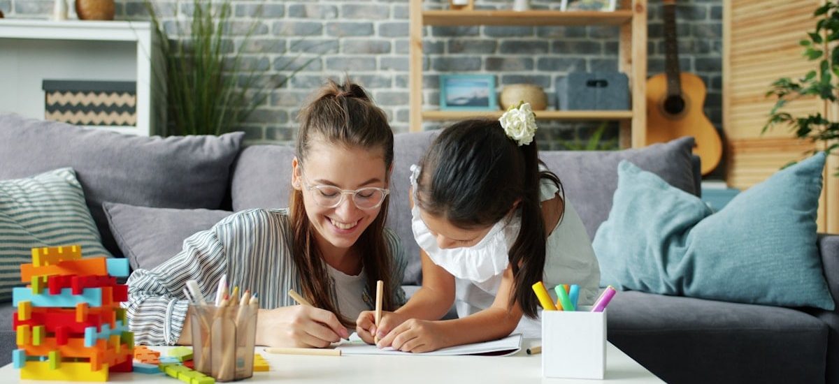 Happy little girl is drawing at home with caring mother using pencils having fun in modern apartment. Creative leisure activity and childhood concept.
