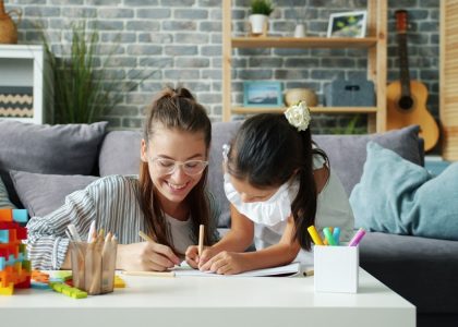 Happy little girl is drawing at home with caring mother using pencils having fun in modern apartment. Creative leisure activity and childhood concept.